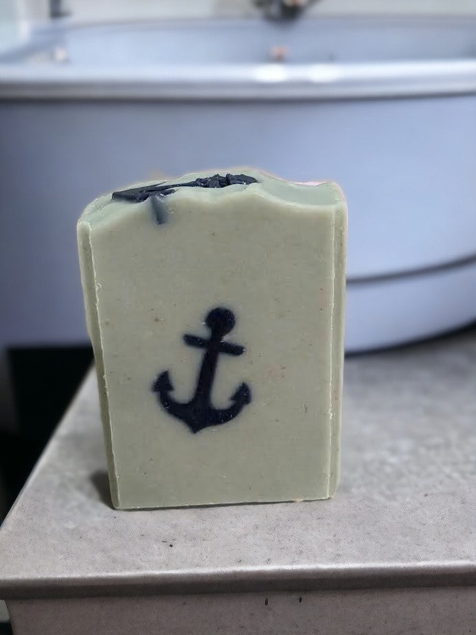 Gentle tallow and goat milk soap featuring simple design of a navy blue anchor against a greenish-blue background. Scented with 'Salty Mariner' the scent is a blend of a blend of lily of the valley, jasmine, leafy greens, pine, and musk, with a splash of fresh rain.  This is a fantastic creamy, bubbly bar soap.