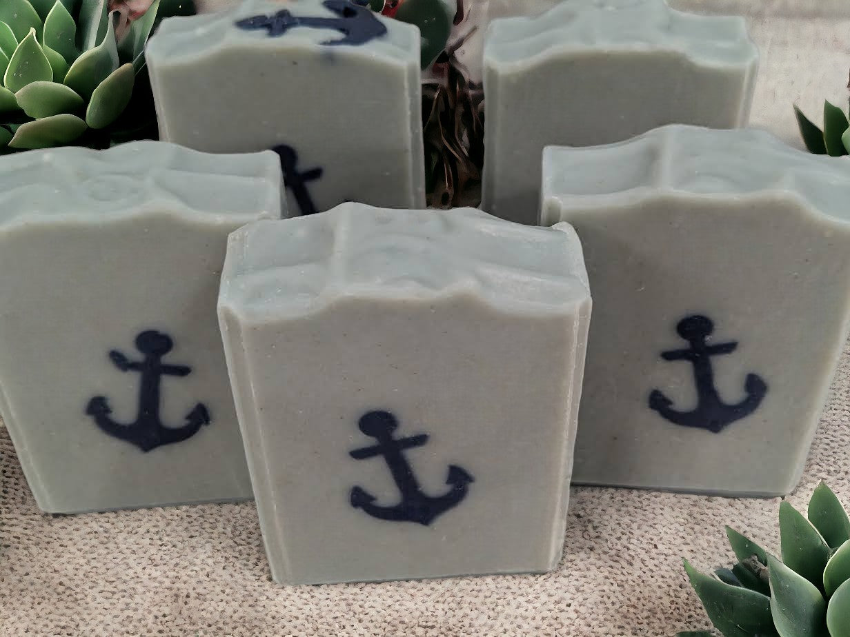 Tallow and goat milk soap featuring simple design of a navy blue anchor against a greenish-blue background. Scented with 'Salty Mariner' the scent is a blend of a blend of lily of the valley, jasmine, leafy greens, pine, and musk, with a splash of fresh rain.  This is a fantastic creamy, bubbly bar soap.